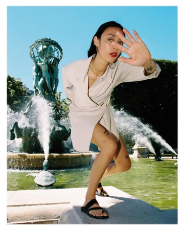 Woman in a short beige dress poses near a fountain and garden on a sunny day, blocking the sun with her hand in an eye-level shot.