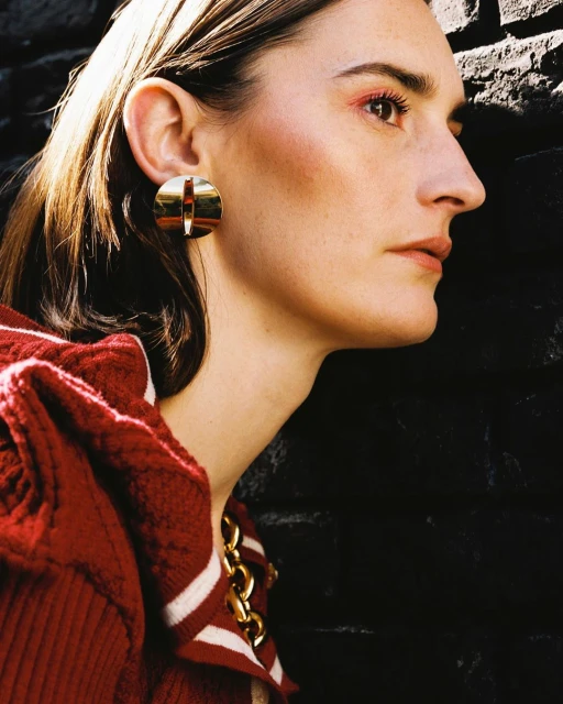 Portrait of a woman in profile with golden disc earrings, wearing a red and white sweater and gold chain necklace, against a dark wall.