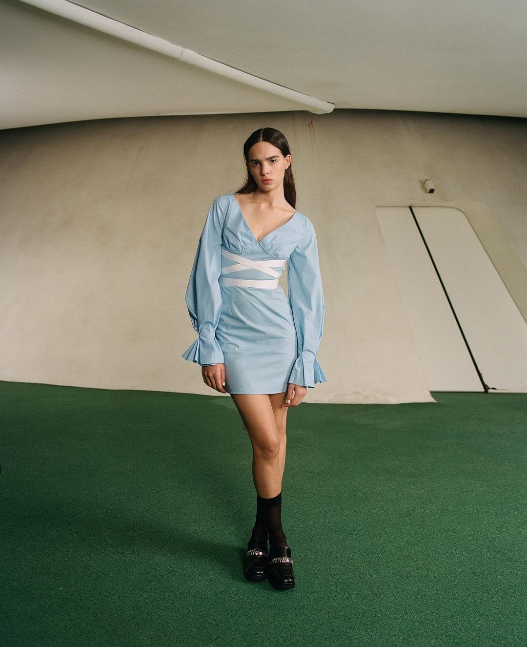 A woman in a light blue dress with puffy sleeves and white cross-straps poses indoors on green flooring, with curved walls behind her.