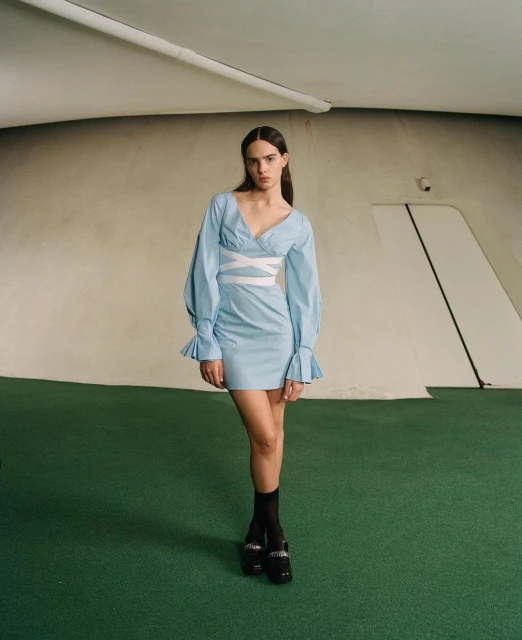 A woman in a light blue dress with puffy sleeves and white cross-straps poses indoors on green flooring, with curved walls behind her.