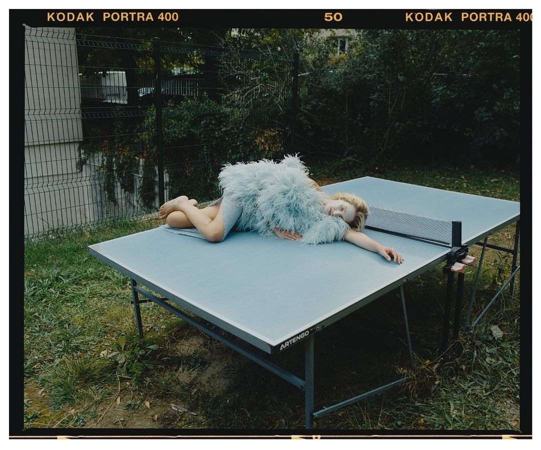 Young woman in a feathered dress rests on a blue ping pong table outside with a chain link fence and greenery in the background.