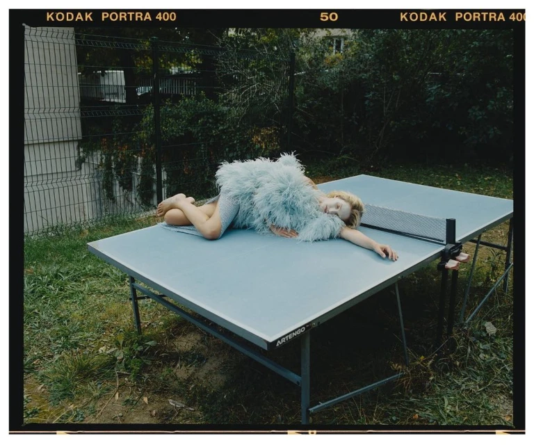 Young woman in a feathered dress rests on a blue ping pong table outside with a chain link fence and greenery in the background.