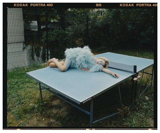 Young woman in a feathered dress rests on a blue ping pong table outside with a chain link fence and greenery in the background.