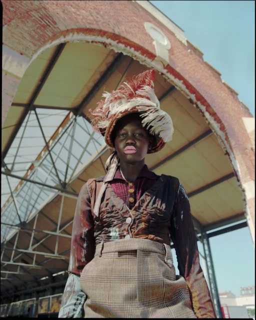 Portrait of a dark-skinned model wearing a feathered hat and vest, posing in front of an old brick building with an arched entrance.