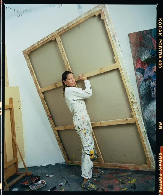 An adult woman in a paint-splattered jumpsuit leans against a large blank canvas inside an artist's studio, holding it in place.