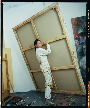 An adult woman in a paint-splattered jumpsuit leans against a large blank canvas inside an artist's studio, holding it in place.