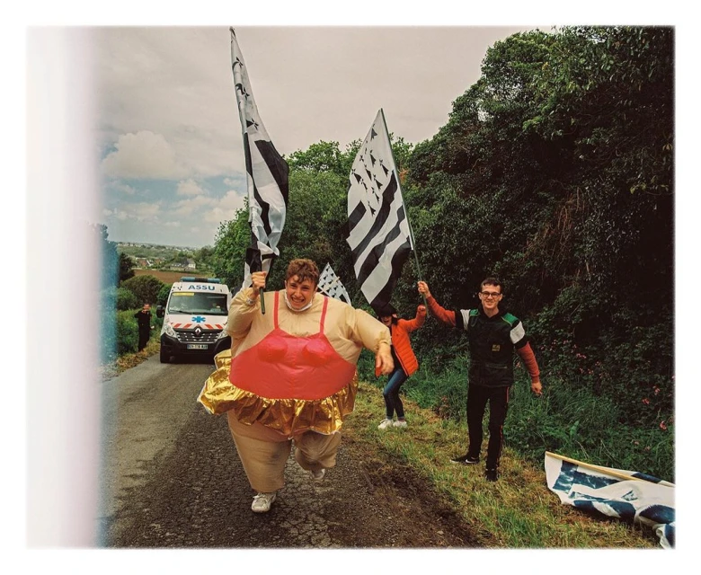 Person in an inflatable sumo wrestler costume runs along a rural road, holding Breton flags, with others cheering, an ambulance in background.