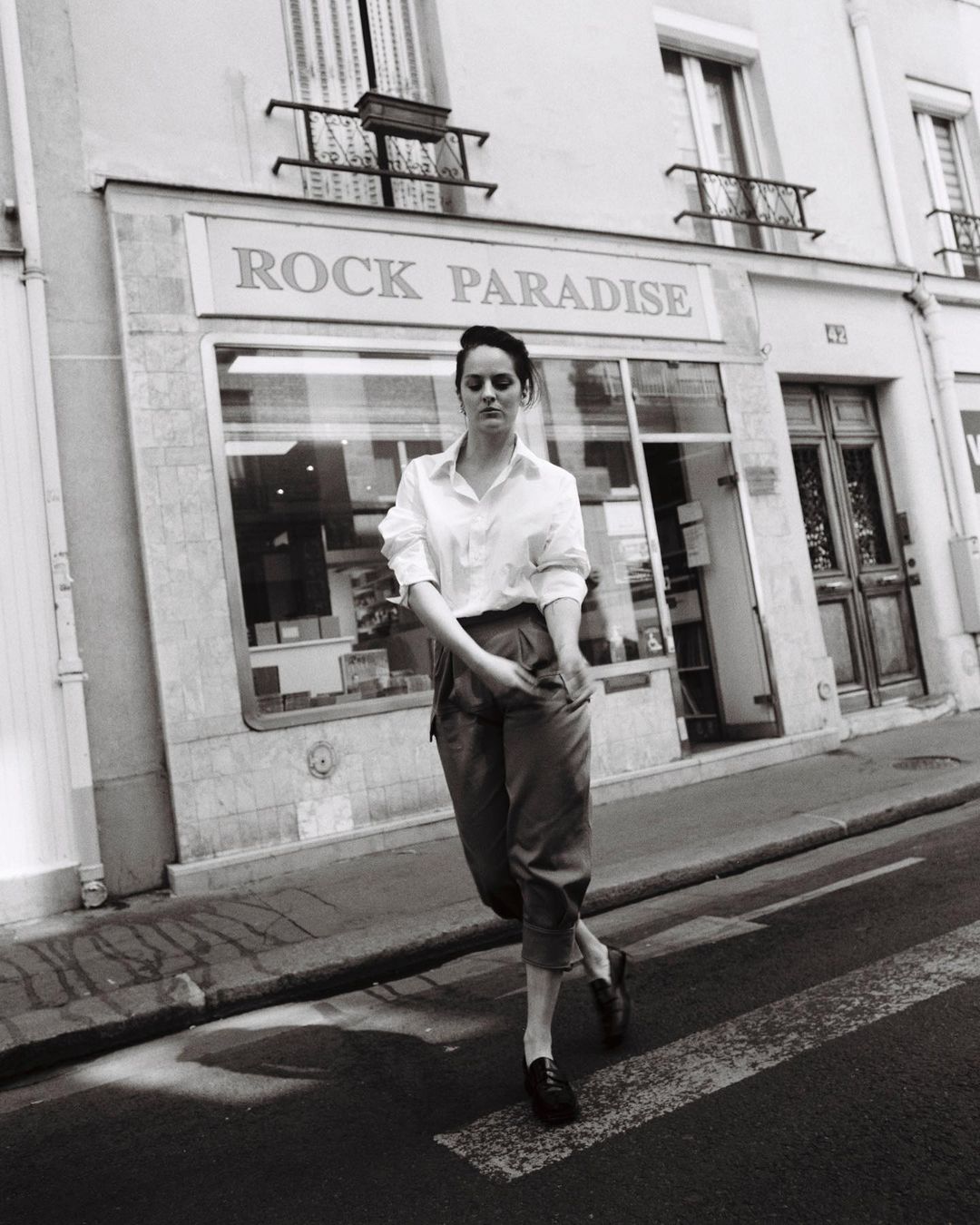 Monochrome portrait of a young woman in button-down shirt and slacks crossing the street in front of Rock Paradise shop.