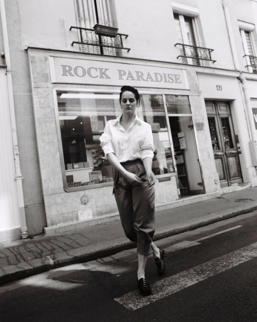 Monochrome portrait of a young woman in button-down shirt and slacks crossing the street in front of Rock Paradise shop.