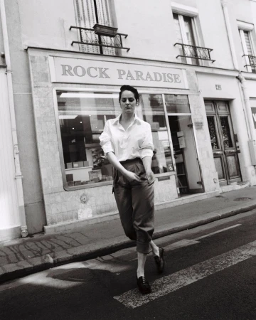 Monochrome portrait of a young woman in button-down shirt and slacks crossing the street in front of Rock Paradise shop.