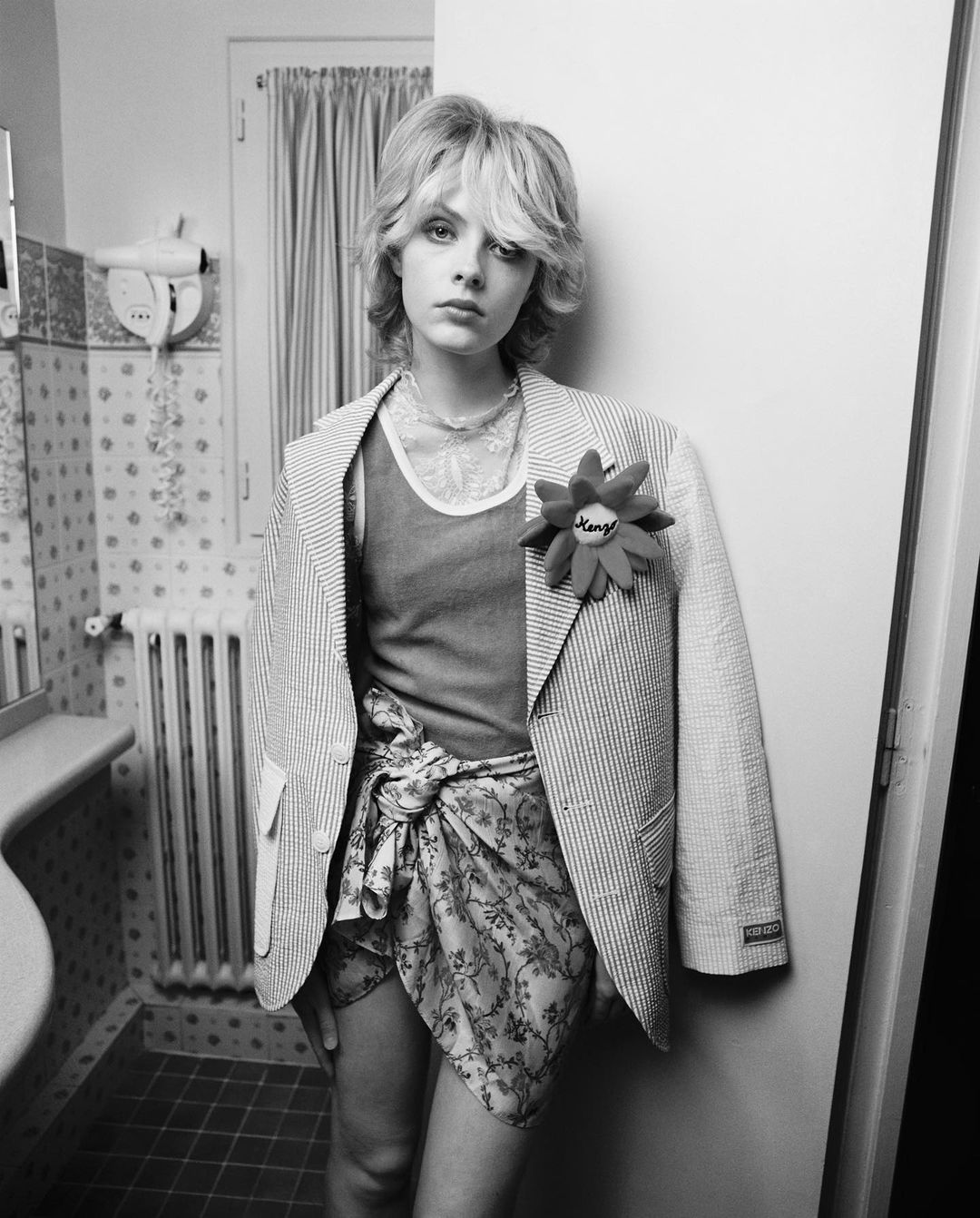 Monochrome portrait of a young adult woman in a blazer and patterned wrap, standing in a bathroom setting with patterned wallpaper.