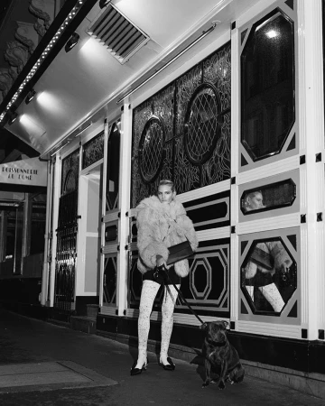 Woman in fur jacket and patterned tights standing on a Paris sidewalk holding a dog leash with a seated dog outside an ornate storefront