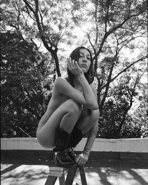A nude young adult woman with tattoos sits on a stepladder wearing cowboy boots outdoors against a backdrop of trees in a black and white photo.