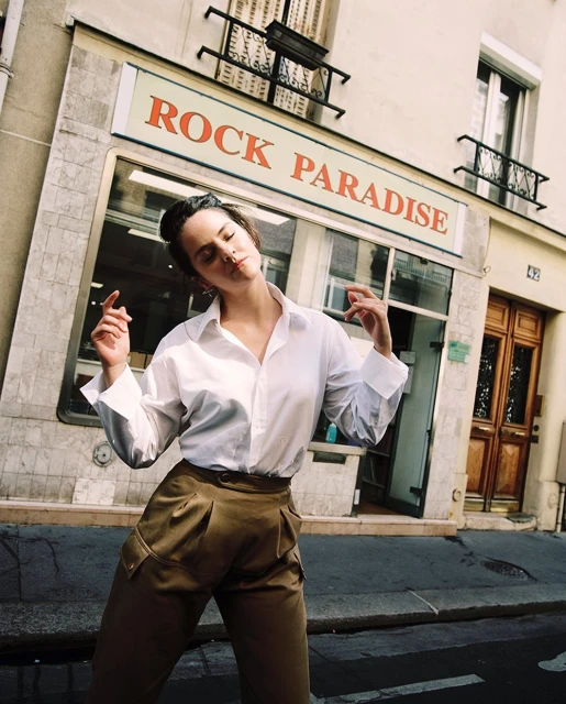 Adult woman posing on a city street, wearing a white button-down shirt and olive-green trousers, near the Rock Paradise store.