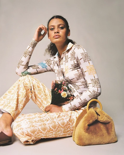 A woman wearing a patterned blouse and pants sits with a bouquet of flowers and a fur bag against a light gray backdrop.