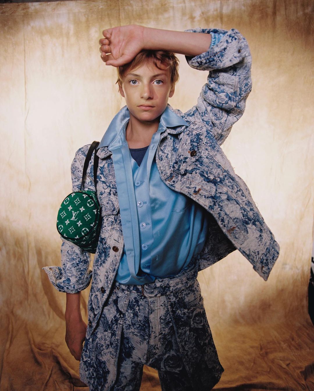 Young male model poses in patterned suit with Louis Vuitton bag against a textured background in a fashion forward, eye-level portrait.