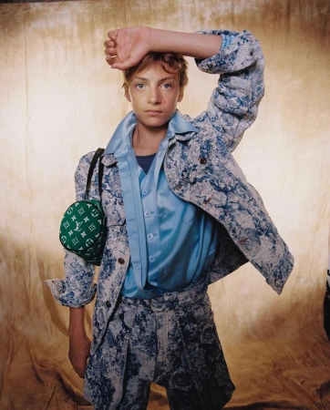 Young male model poses in patterned suit with Louis Vuitton bag against a textured background in a fashion forward, eye-level portrait.