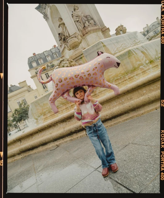 A girl in jeans stands in front of a stone fountain holding a large pink and gold leopard balloon on her shoulders in urban environment.