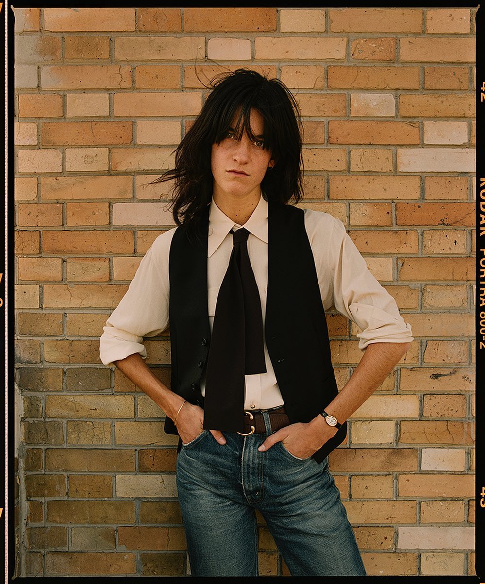 An androgynous person with long dark hair, wearing a black vest and blue jeans, poses in front of a brick wall in a portrait.