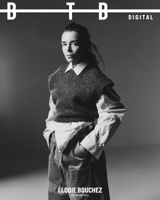Monochrome portrait of a young woman in a collared shirt and vest, standing in front of a grey backdrop. Magazine cover.