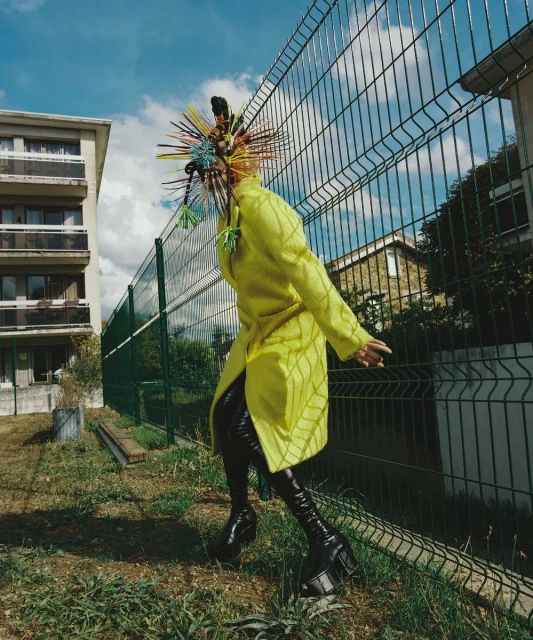 Person in yellow coat and avant-garde headpiece stands near a chain-link fence and buildings on a grassy lawn under a blue sky with clouds.