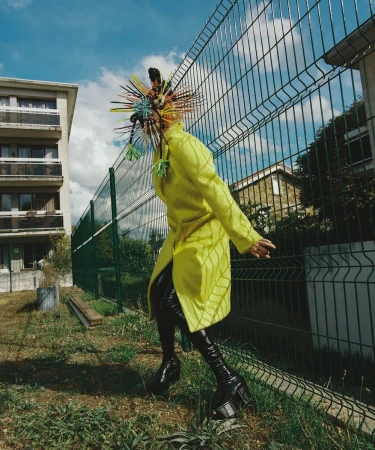 Person in yellow coat and avant-garde headpiece stands near a chain-link fence and buildings on a grassy lawn under a blue sky with clouds.