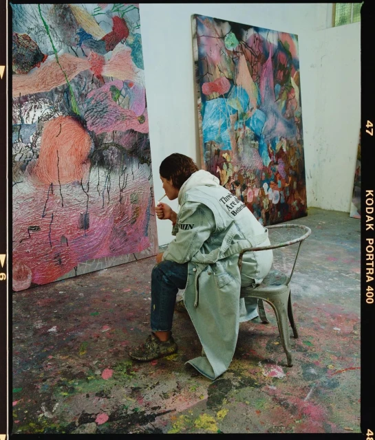 Artist in light blue denim jacket with lettering sits on metal chair in art studio with two large abstract paintings in the background.