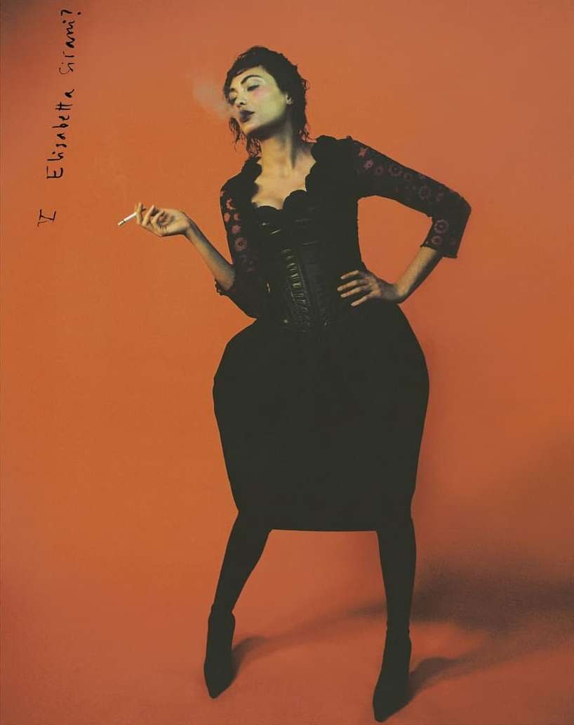 A woman in a dark dress and heels stands against a red-orange backdrop, smoking a cigarette, in a fashion photography portrait.