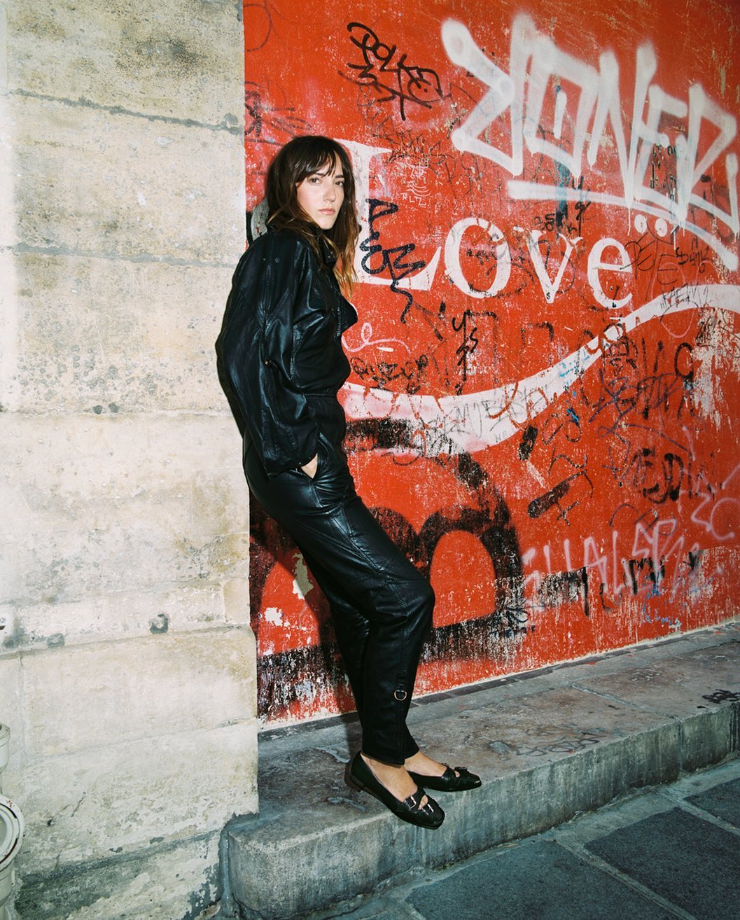 Young woman in black leather outfit poses against graffiti-covered wall in urban setting, showcasing fashion-forward style and texture.