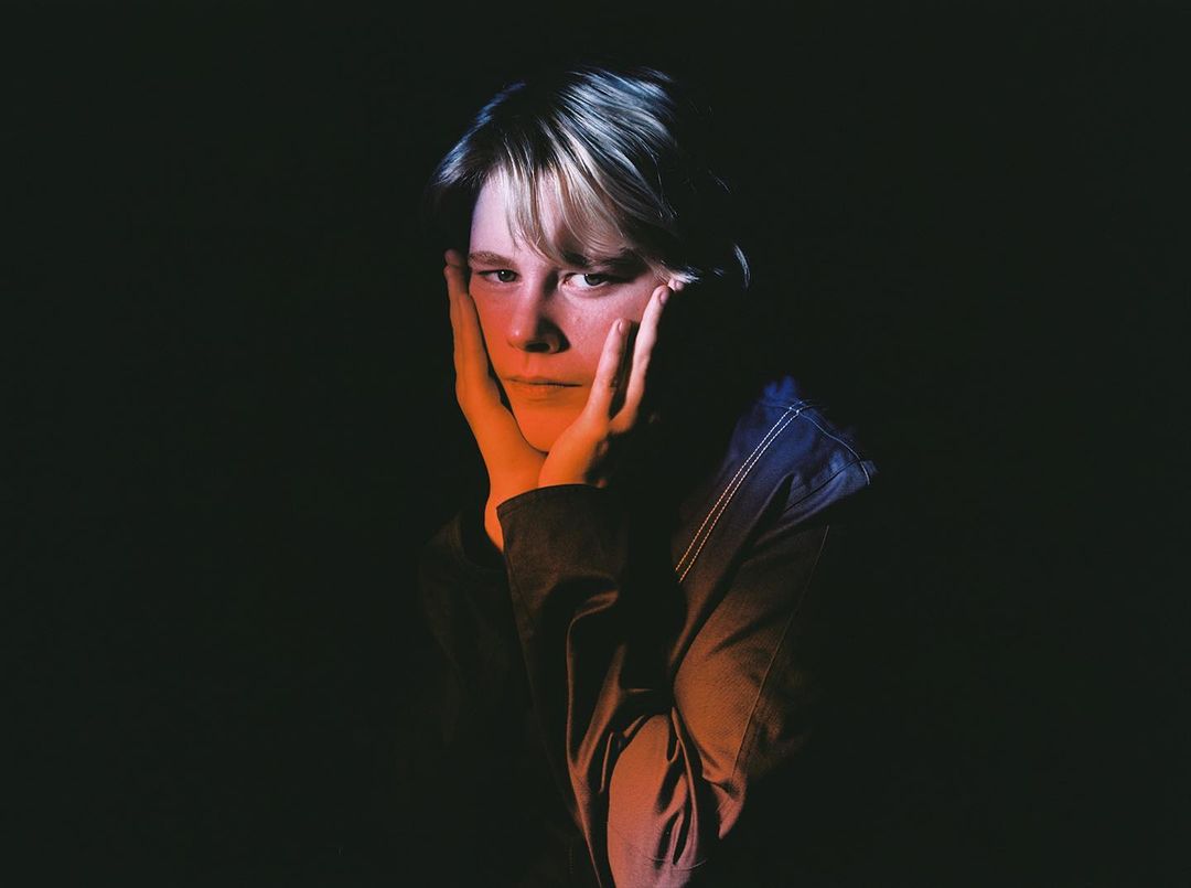 Close-up portrait of a person with light hair with hands on their face against a dark background with orange and blue rim lighting.