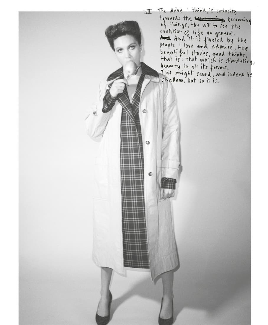 Adult female in plaid dress and beige trench coat holds sphere in an indoor photo studio with script handwriting in the background.