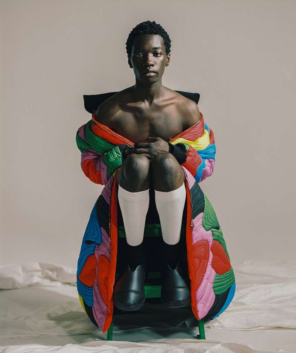Striking studio portrait of an African male model in a vibrant quilted jacket, seated on a chair, with a neutral backdrop and soft lighting.