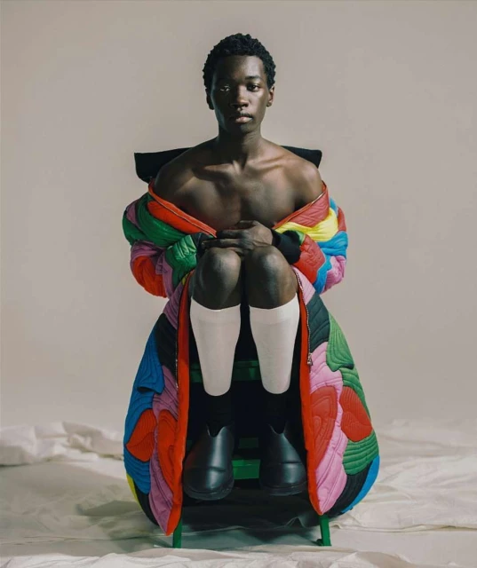 Striking studio portrait of an African male model in a vibrant quilted jacket, seated on a chair, with a neutral backdrop and soft lighting.