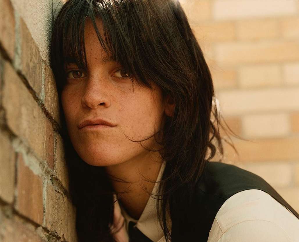 Portrait of an adult woman with short dark hair leaning against a brick wall, wearing a white shirt and dark vest.
