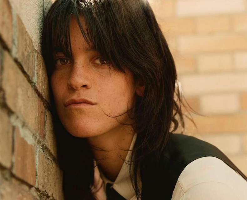 Portrait of an adult woman with short dark hair leaning against a brick wall, wearing a white shirt and dark vest.