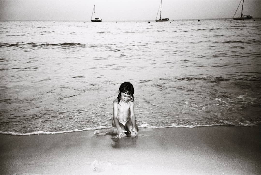 A child sits on a sandy beach near the water's edge, with several sailboats visible in the distance on the ocean in a black-and-white photo.