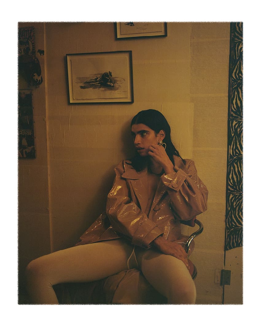 Non-binary person sitting on a chair in a vintage interior, wearing a shiny pink jacket and off-white tights, posing for a portrait.