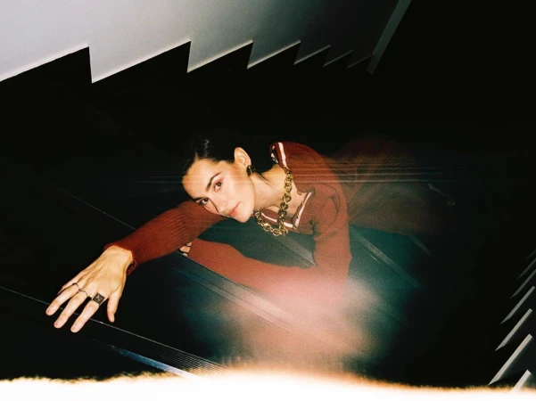 Brunette woman in a maroon outfit and gold jewelry poses on a staircase with dark shadows and white risers.