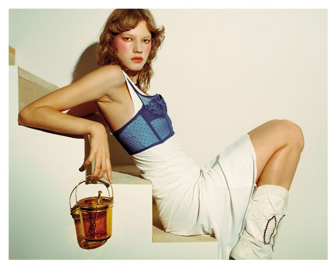 Model in blue corset and white skirt poses on steps with transparent amber bucket bag in a studio fashion editorial, facing camera.