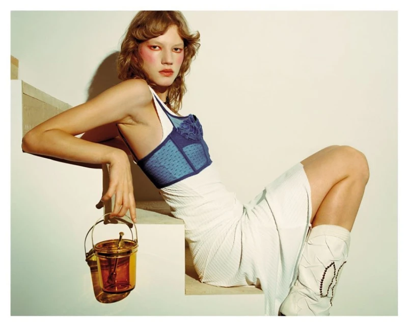 Model in blue corset and white skirt poses on steps with transparent amber bucket bag in a studio fashion editorial, facing camera.