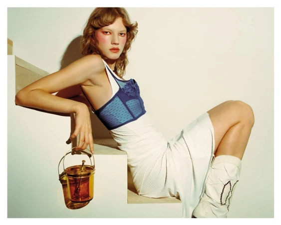 Model in blue corset and white skirt poses on steps with transparent amber bucket bag in a studio fashion editorial, facing camera.