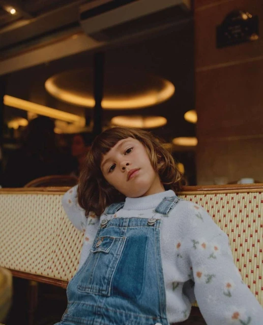 Young child in blue denim overalls and a floral sweater sits on a woven chair in a warm-lit cafe with a contemplative gaze.