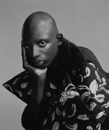 Portrait of a bald black man wearing a dark patterned jacket, resting his chin on his hand against a plain studio background.