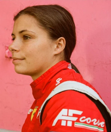 Profile portrait of a woman in a red racing suit against a pink background. She is fair-skinned with dark hair in a ponytail.