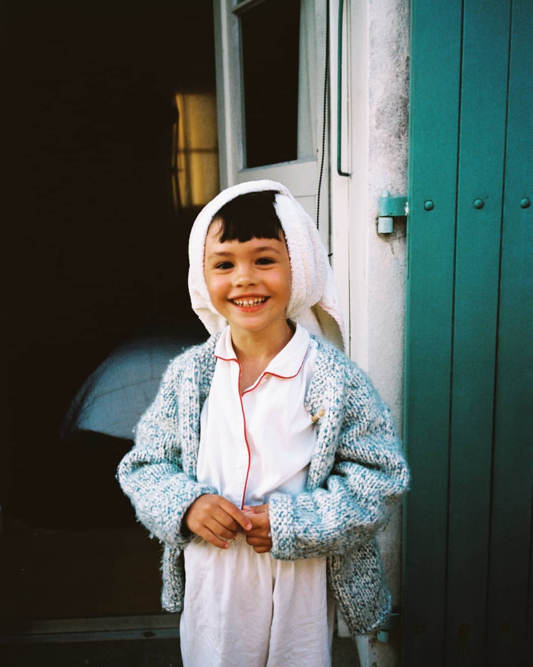 Smiling young girl wearing pajamas, a cardigan, and a towel on her head, standing in front of a teal door on a sunny day.