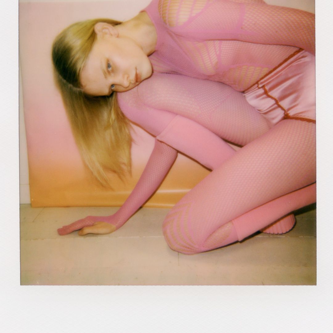 A blonde woman in a pink fishnet top and shorts kneels on a light surface with her head resting on her knee in a studio shot.