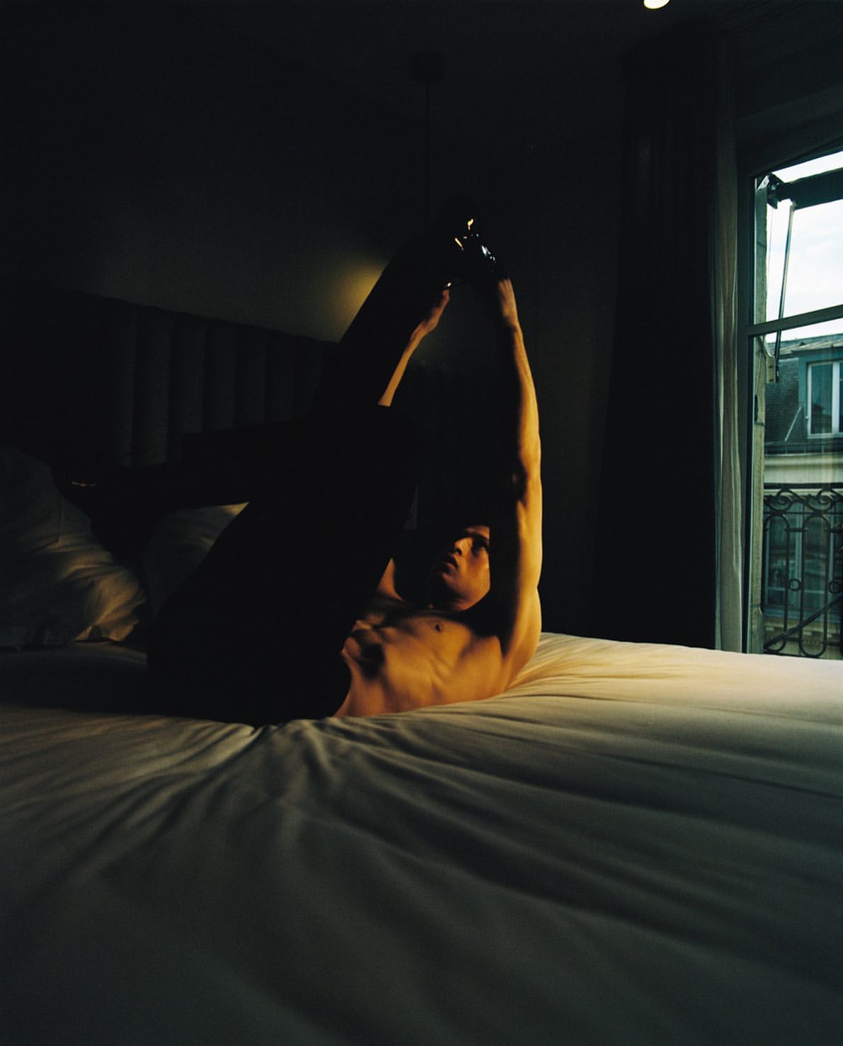Man in arched pose on bed in dark room with window. Nude torso and raised arms, room lit with dim natural light.
