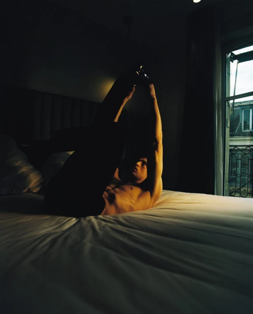 Man in arched pose on bed in dark room with window. Nude torso and raised arms, room lit with dim natural light.