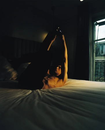 Man in arched pose on bed in dark room with window. Nude torso and raised arms, room lit with dim natural light.