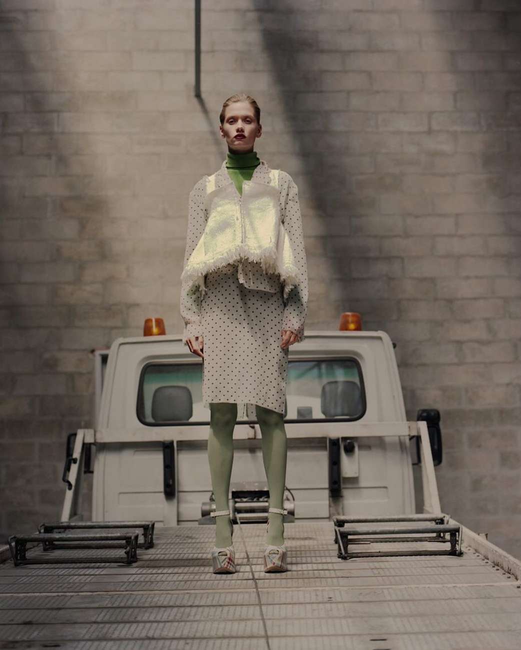 A fair-skinned fashion model poses in a dotted blouse and skirt, green tights, and platform heels, standing on the back of a truck.